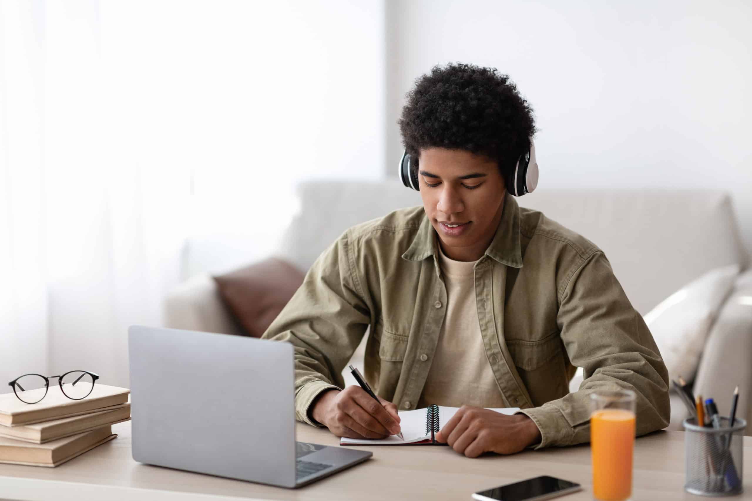 Studying from home concept. African American teenage guy in headphones writing down info during online lecture indoors, free space. Black male student participating in webinar or internet conference